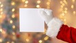 © Viktoriia - Santa's hand holding a blank piece of paper. On festive holiday background. Free space for text. New Year postcard. Santa Claus wearing white gloves, closeup.