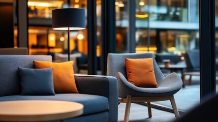 Sticker - Modern lounge area with gray sofa and armchair featuring orange and gray cushions, illuminated by stylish floor lamp, reflecting warm interior ambiance in glass windows.