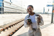 © Jelena Stanojkovic - Smiling African-American woman enjoying the moment at train station, holding coffee and phone. Its relaxed appearance and casual style indicate readiness for a trip or a daily adventure.