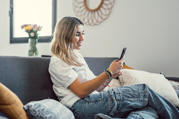 Wall Mural - Happy pleasant millennial girl relaxing on comfortable couch, holding smartphone in hands. Smiling young lady chatting in social networks, watching funny videos, using mobile applications at home..