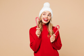 Canvas Print - Merry young woman wear red warm cozy sweater hat posing hold in hand candy cane stick look aside isolated on plain pastel light beige background. Happy New Year celebration Christmas holiday concept.