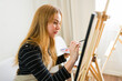 © AntonioDiaz - Focused young female artist painting on canvas with easel, palette, and brush in her studio