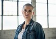 © Running opossum - Portrait of confident woman with shaved head wearing denim jacket standing in front of large windows in industrial loft