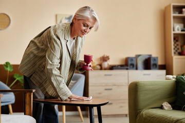 Side view portrait of elegant mature woman enjoying Spring cleaning at home and dusting coffee table in cozy living room copy space