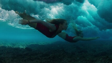 Wall Mural - Two women in sensual red suits dive under the breaking wave and keep gliding underwater in the foam.