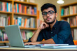 © GustavsMD - A thoughtful young man wearing glasses sits in a library, surrounded by books, working on a laptop. Perfect for concepts of education, focus, and academic life