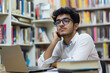 © GustavsMD - Thoughtful young male student with glasses working on a laptop in a library, surrounded by books