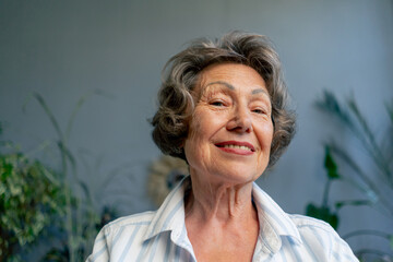 close-up of a very elderly gray-haired grandmother in a light blouse with a slight smile sitting against the background of a blue wall with large indoor plants