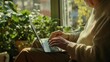 © nabila - Elderly Person Working on Laptop Surrounded by Lush Green Plants in a Cozy Indoor Setting During a Bright Sunny Day, Embracing Nature and Technology Harmony