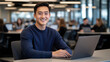 © ERiK - A young professional, smiling and sitting at a desk, works on a laptop in a vibrant, modern office setting with colleagues in the background.