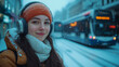 © sommersby - A young woman smiles while wearing headphones and a warm hat, standing in the snow as she waits for a bus in a busy urban area