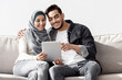 © Prostock-studio - Joyful muslim family sitting on couch in new apartment, using pad, copy space. Happy middle-eastern young man and woman in hijab buying furniture for house, unpacking stuff after moving