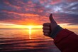 © Aleksandra - thumbs up at sunset on the beach with ocean reflection and vibrant sky