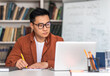 © Prostock-studio - E-Teaching. Korean Middle Aged Man Teacher Using Laptop And Taking Notes Preparing For Virtual Lecture Sitting At Desk In Modern Classroom. Internet Technology And Education. Selective Focus