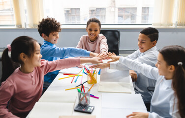 Teamwok And Children Concept. Diverse excited group of emotional happy junior school kids sitting at desk in classroom and putting hands together, having fun, studying or playing game