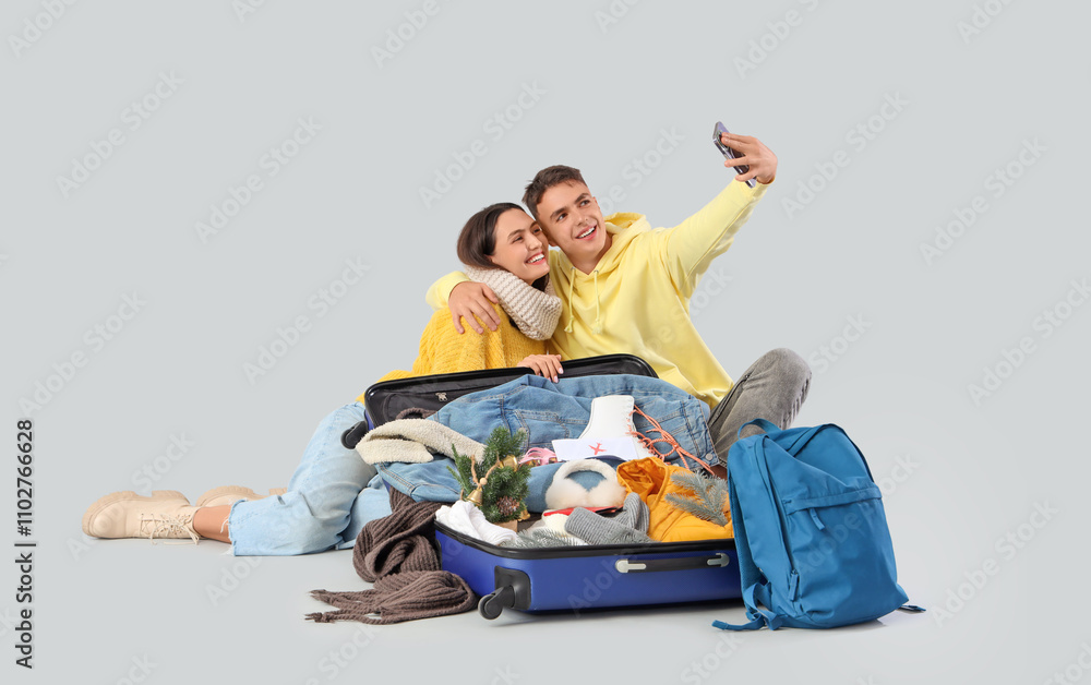 Cute young couple in warm clothes taking selfie with open suitcase on grey background. Winter vacation concept
