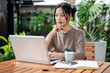© bongkarn - An attractive Asian woman sitting at an outdoor table of a cafe, working remotely.