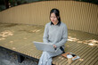 © bongkarn - A charming Asian woman works remotely outdoors, sitting on a park bench with her laptop.
