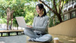 © bongkarn - A charming Asian woman works remotely outdoors, sitting on a park bench with her laptop.
