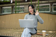 © bongkarn - A charming Asian woman works remotely outdoors, engaged in an online meeting through her laptop.