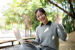 © bongkarn - A beautiful Asian woman sits on a park bench with her laptop, smiles warmly and waves at the camera.