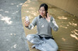 © bongkarn - A charming woman sits on a park bench, talking on a video call with her friend through her phone.