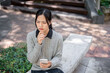 © bongkarn - A thoughtful Asian woman sits outdoors, holding her smartphone in her hand, deep in thought.