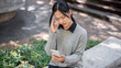 © bongkarn - A stressed Asian woman receiving unexpected bad news through a message while sitting outdoors.