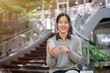 © bongkarn - A happy Asian woman sits outdoors on a bright day, holding a smartphone and smiling at the camera.