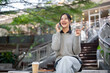© bongkarn - A cheerful, smiling Asian woman enjoys talking on the phone while sitting outdoors in the city.