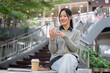 © bongkarn - A beautiful, happy Asian woman sits outdoors on a bright day, using her smartphone with a smile.