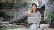 © bongkarn - A charming Asian woman enjoys a hot drink while working on her laptop outdoors.