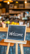 © Anastasia - A minimalistic welcome sign on an easel stands out against a blurred café background, inviting guests with a warm and friendly atmosphere