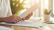 © Alexander - A close-up of a person flipping through a stack of documents on a wooden desk in a brightly lit office. The focus is on their hands, the clean white papers, and the warm glow of na