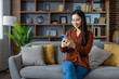 © Liubomir - Asian woman sits on sofa using smartphone, smiling while engaged in online activity. Modern home setting with stylish decor and comfortable seating creates relaxing environment. Technology in everyday