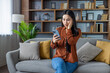 © Liubomir - Surprised asian woman using smartphone at home. Sitting on sofa, reading unexpected message on phone, hand to face in disbelief. Casual setting with home decor around.