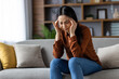 © Liubomir - Asian woman having headache, sitting on couch at home. Pressing temples with fingers, looking stressed and uncomfortable. Casual clothing, indoor setting, feeling tension and fatigue.