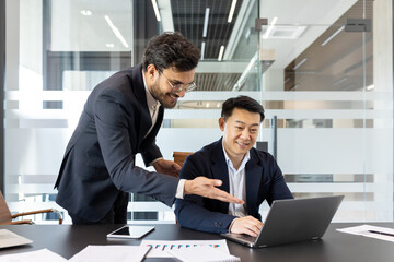 Wall Mural - Businessmen collaborating in office. Focused team discussing project at laptop. One standing, one sitting, with smartphone and documents on table. Professional teamwork concept.