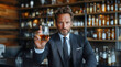 © Liuda - Confident man wearing a dark suit. Whiskey glass raised in hand. Shelves filled with bottles in the background. Stylish bar setting. Sophisticated atmosphere. Focused and poised expression