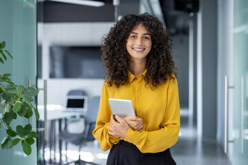 confident woman in office holding tablet, smiling warmly. curly hair, yellow blouse, modern workspac
