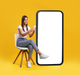 Cheerful smiling female sitting on chair near big cell phone with empty white screen, using mobile device, checking new cool app on orange studio background, mock up for website or application design