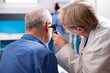 © DC Studio - Retired man seated in hospital room undergoes hearing test as senior female ent specialist performs medical examination with otoscope. Elderly doctor checking ear of male pensioner patient in clinic.