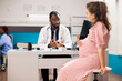 © DC Studio - Expectant mother talks to african american obstetrician about her pregnancy progress during medical checkup. Friendly doctor listening to pregnant caucasian woman while seated at clinic office desk.