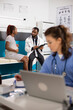 © DC Studio - Physician in lab coat gives prenatal advice to pregnant lady while nurse sits at nearby desk reviewing medical files. Female healthcare worker reads patient records while doctor consults with patient