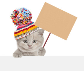 Fototapete - Cute kitten wearing a warm hat with pompon looking above blank banner and showing blank placard on wooden stick. isolated on white background
