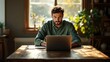 © LittleBird919 - A man in a green shirt works on a laptop by a window.