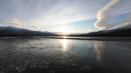 Wall Mural - A serene view showcases the sun setting over an icy river, reflecting warm hues on the frozen surface. Snow-capped mountains frame the horizon, creating a peaceful winter landscape.