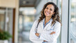 © TimosBlickfang - Confident female doctor with a stethoscope smiles warmly, standing in a bright hospital corridor with arms crossed.