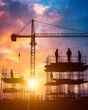 © Thanaseth - Silhouetted workers on a construction site, with the warm hues of sunset reflecting off building materials and cranes