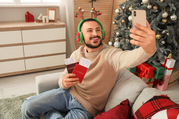 Sticker - Young man taking selfie with passports on sofa in living room. Christmas travel concept